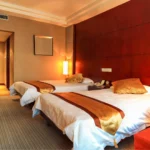 A sophisticated hotel suite bedroom showcasing a custom decorative headboard accent wall crafted from Premium Red Grandis™ quarter cut wood veneer by theShaper, displaying its signature elegant rosy-pink hue and perfectly straight linear grain.